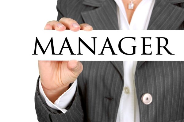 Gendern, ein Leitfaden für Unternehmen: ein Frauenoberkörper, gekleidet in weißer Bluse und grauem Jackett, in einer Hand hält die Frau ein Schild mit der Aufschrift "Manager"