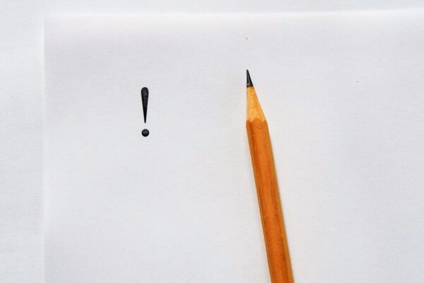 Ausrufezeichen verwenden: weißer Hintergrund, darauf ein weißer Papierbogen, links steht ein schwarzes Ausrufezeichen, rechts liegt ein Bleistift