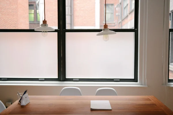 Satzzeichen richtig setzen: brauner Schreibtisch mit Notizblock und Stiftehalter mit schwarzem und weißem Stift, vier weiße Stühle, dahinter eine weiße Wand, zwei Fenster und zwei von der Decke hängende Lampen