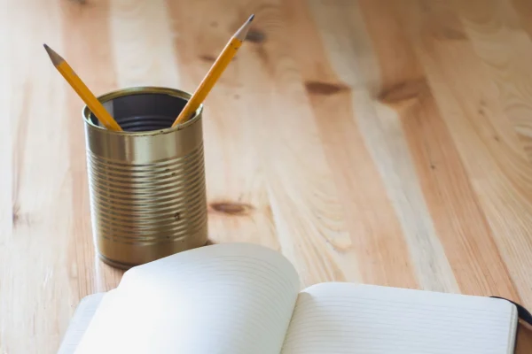 nicht abschließende Aufzählungen: Schreibtisch mit geöffneten Notizbuch und Blechdose, aus der zwei Bleistifte ragen