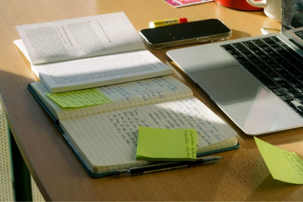 Werbetexte selbst überarbeiten: Schreibtisch mit geöffnetem Notebook, Smartphone, geöffnetem Buch, geöffnetem und beschrifteten Notizbuch und mehreren beschrifteten Notizzetteln